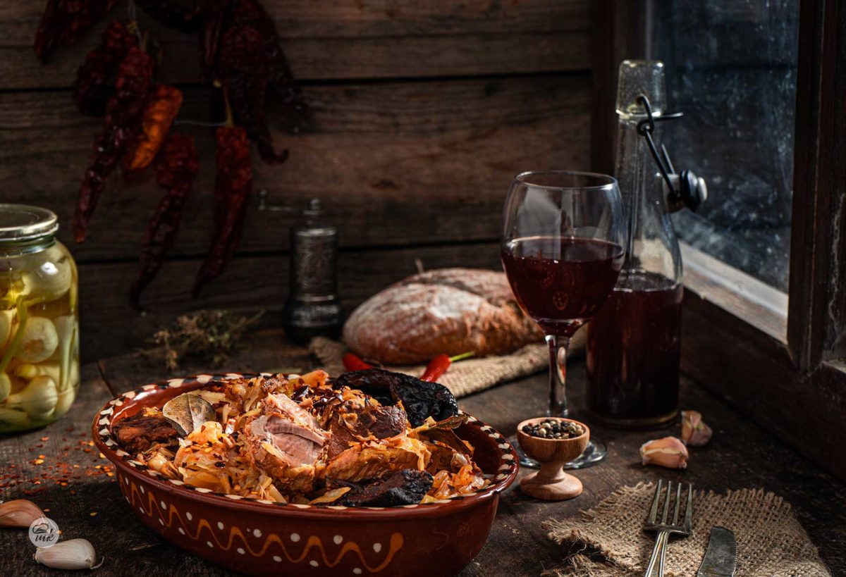 Кисело зеле със свинско и пушена сланина, сервирано в глинен съд, на заден фон с хляб и червено вино, снимано отстрани