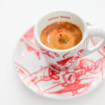 Чаша кафе в бяла чашка с червени мотиви върху чинийка, снимано под ъгъл