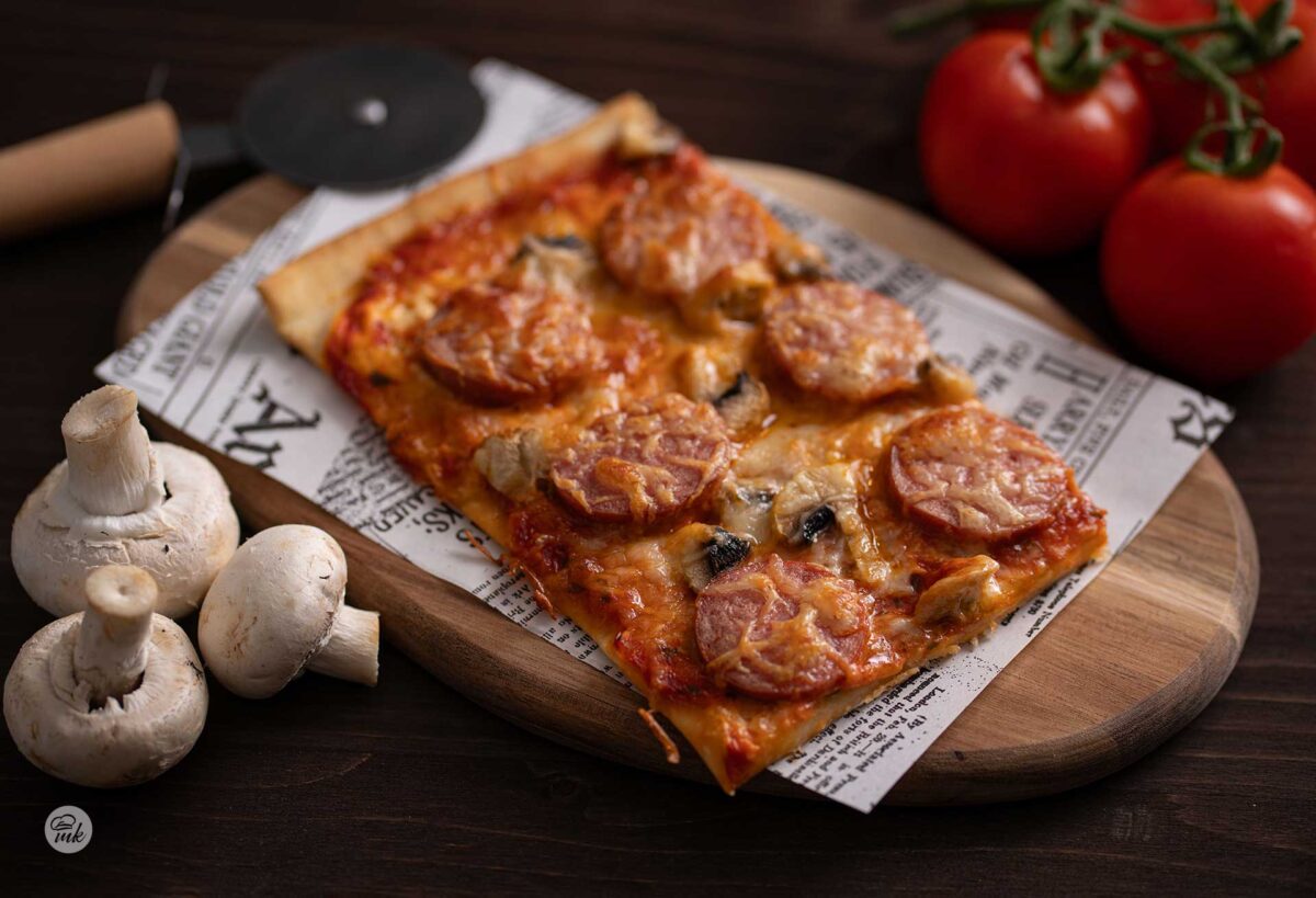 Правоъгълно парче пица с наденица и гъби върху дъска, покрита с хартия