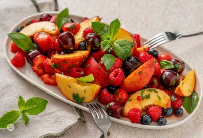 Плодова салата от праскови, череши, ягоди, малини и боровинки в бял, порцеланов поднос, гарнирана с листенца прясна мента.